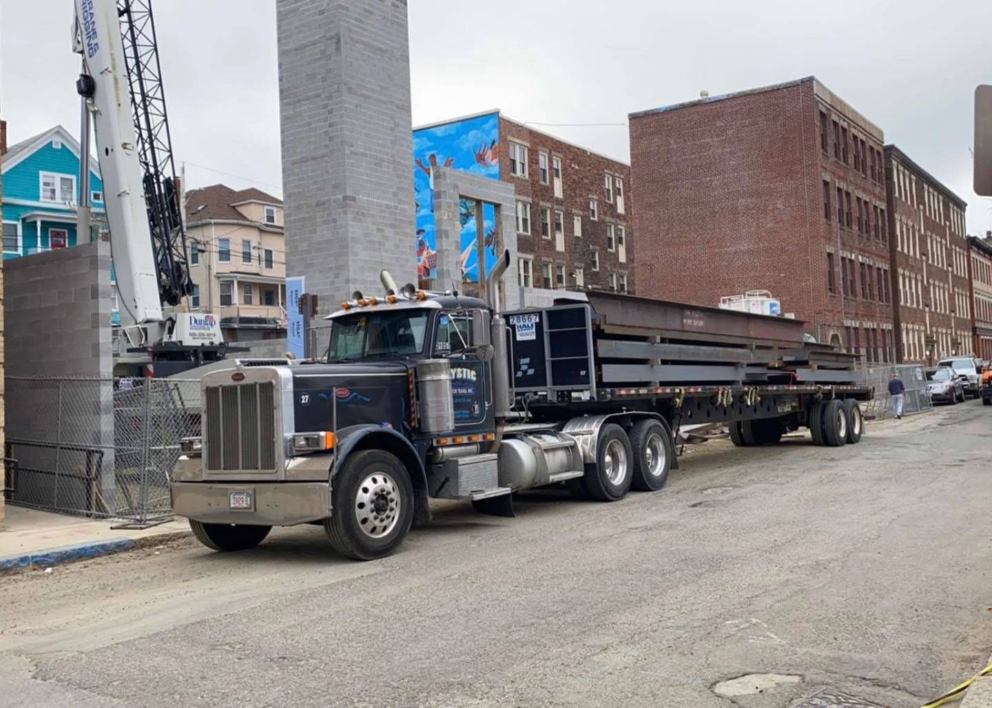 Black flatbed semi-truck delivering heavy structural steel beams to an urban construction site with a crane in the background.